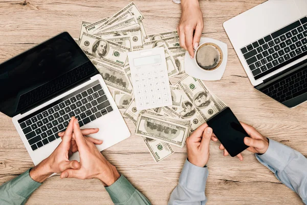 Top view of business partners with laptops and smartphone by calculator on dollar banknotes on table, cropped view — Stock Photo