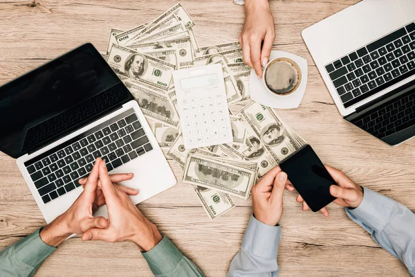 Vista dall'alto dei partner commerciali con computer portatili e smartphone tramite calcolatrice sulle banconote in dollari sul tavolo, vista ritagliata — Foto stock