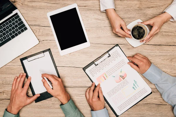 Ansicht von Geschäftspartnern mit Papierkram, digitalen Geräten und Kaffee auf dem Tisch, beschnittene Ansicht — Stock Photo