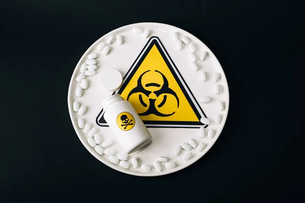 Vue du dessus du panneau de danger biologique sur l'assiette avec des pilules et bouteille avec lettrage poison isolé sur noir — Photo de stock