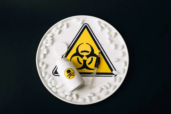 Top view of pills, syringe and jar on plate with biohazard sign isolated on black — Stock Photo