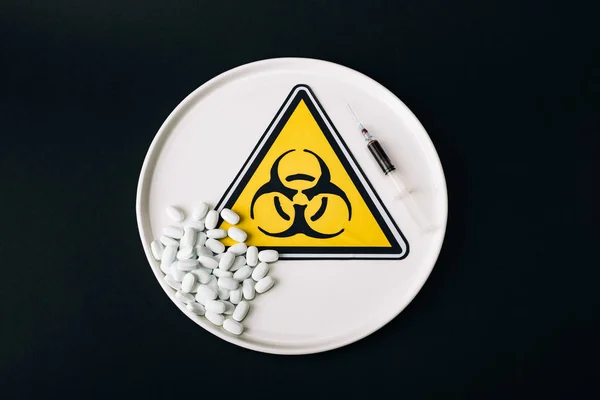 Top view of pills and syringe on plate with biohazard sign isolated on black — Stock Photo