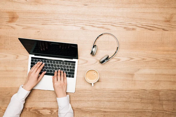 Vista dall'alto della donna che utilizza laptop da caffè e cuffie su tavolo in legno, vista ritagliata — Foto stock