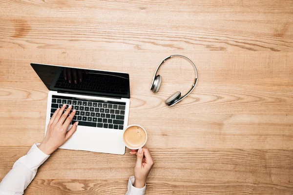 Top view of woman using laptop and drinking coffee by headphones on wooden table, cropped view — Stock Photo