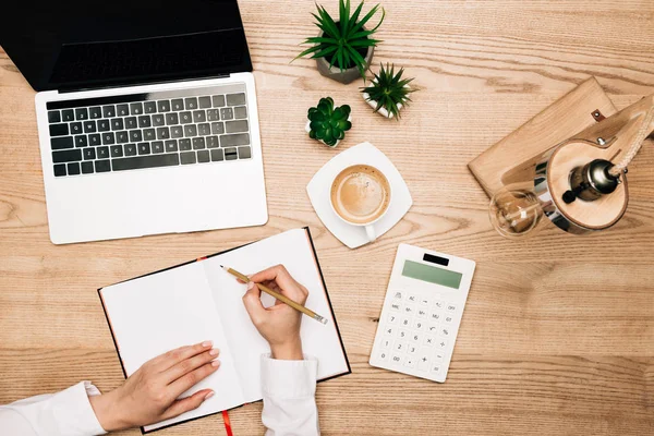 Visão superior da mulher de negócios escrevendo em notebook por calculadora, laptop e café na mesa — Fotografia de Stock
