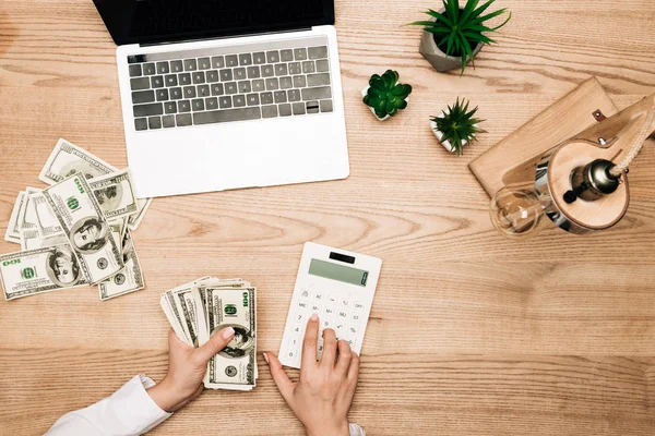 Ansicht von oben: Geschäftsfrau hält Dollarnoten in der Hand, während sie Taschenrechner mit Laptop auf dem Tisch benutzt, beschnittene Ansicht — Stockfoto