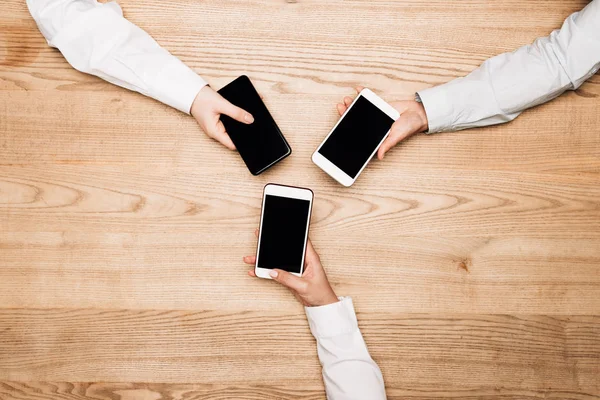 Vista superior de gente de negocios sosteniendo teléfonos inteligentes en la mesa de madera, vista recortada - foto de stock