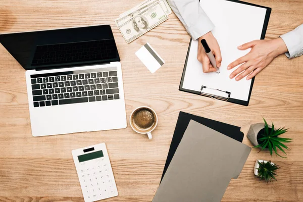 Top view of businessman writing on clipboard with laptop, money and credit card on table — Stock Photo