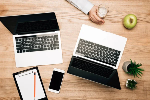 Vista superior del hombre de negocios sosteniendo un vaso de agua por computadoras portátiles, teléfono inteligente y portapapeles en la mesa - foto de stock