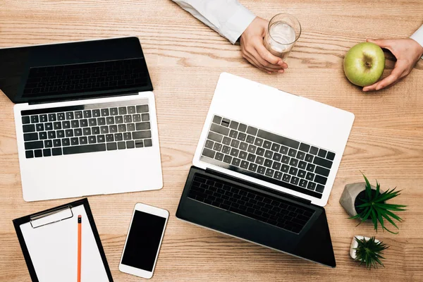 Vista superior del hombre de negocios sosteniendo manzana y vaso de agua por computadoras portátiles y portapapeles en la mesa - foto de stock