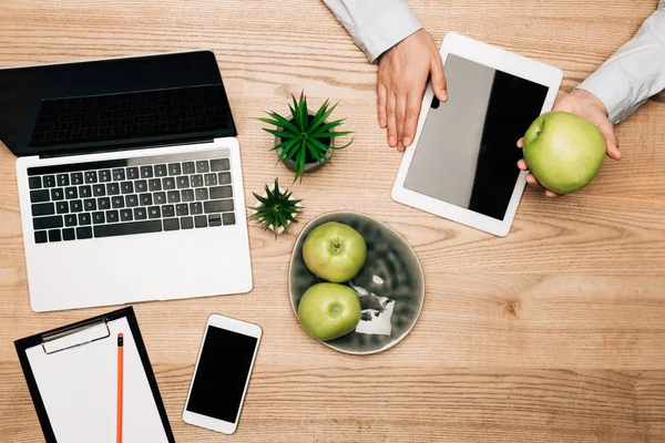 Вид сверху бизнесмена, держащего яблоко рядом с цифровым планшетом, ноутбуком и смартфоном на столе — стоковое фото