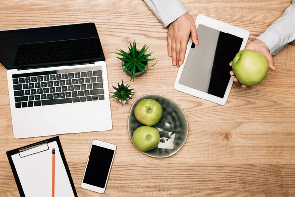 Vista superior del hombre de negocios que sostiene la manzana junto a la tableta digital, portátil y teléfono inteligente en la mesa - foto de stock