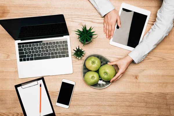 Vista superior do homem de negócios com gadgets digitais segurando maçã na mesa de madeira, vista cortada — Fotografia de Stock