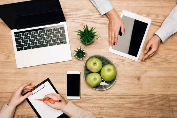 Top view of colleagues with digital devices and clipboard on table, cropped view — Stock Photo