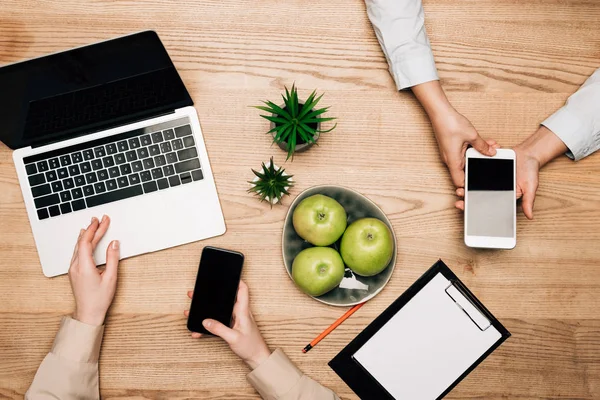 Ansicht von Geschäftsleuten, die Smartphones und Laptops mit Äpfeln und Klemmbrett auf dem Tisch benutzen — Stockfoto