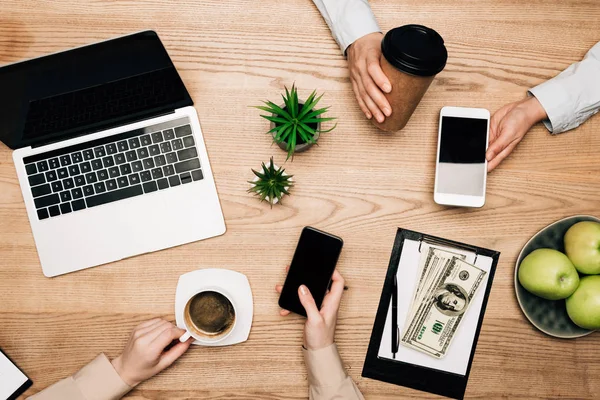 Top view of business partners drinking coffee and using smartphones by laptop and money on table — Stock Photo