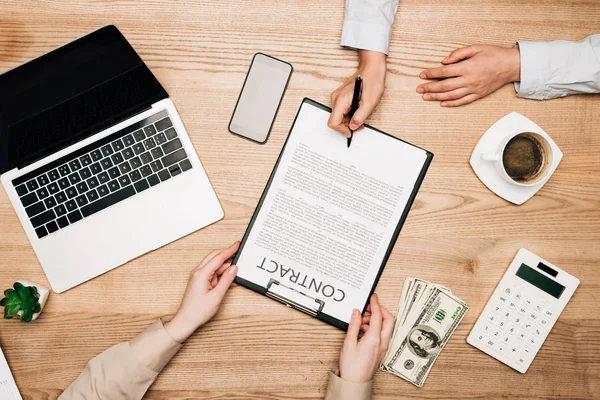 Top view of business partners sign contract by laptop, dollar banknotes and coffee on table, cropped view — Stock Photo