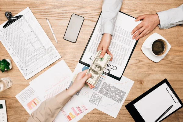 Vista superior del hombre de negocios dando billetes de dólar a su colega con contrato y papeleo en la mesa - foto de stock