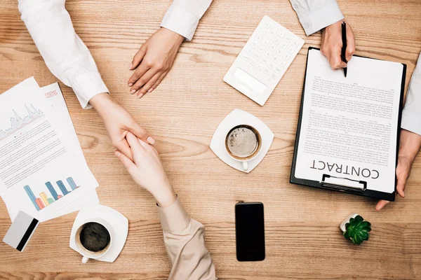 Вид сверху на деловых партнеров, пожимающих руки возле контракта, кофе и кредитная карта на столе — стоковое фото