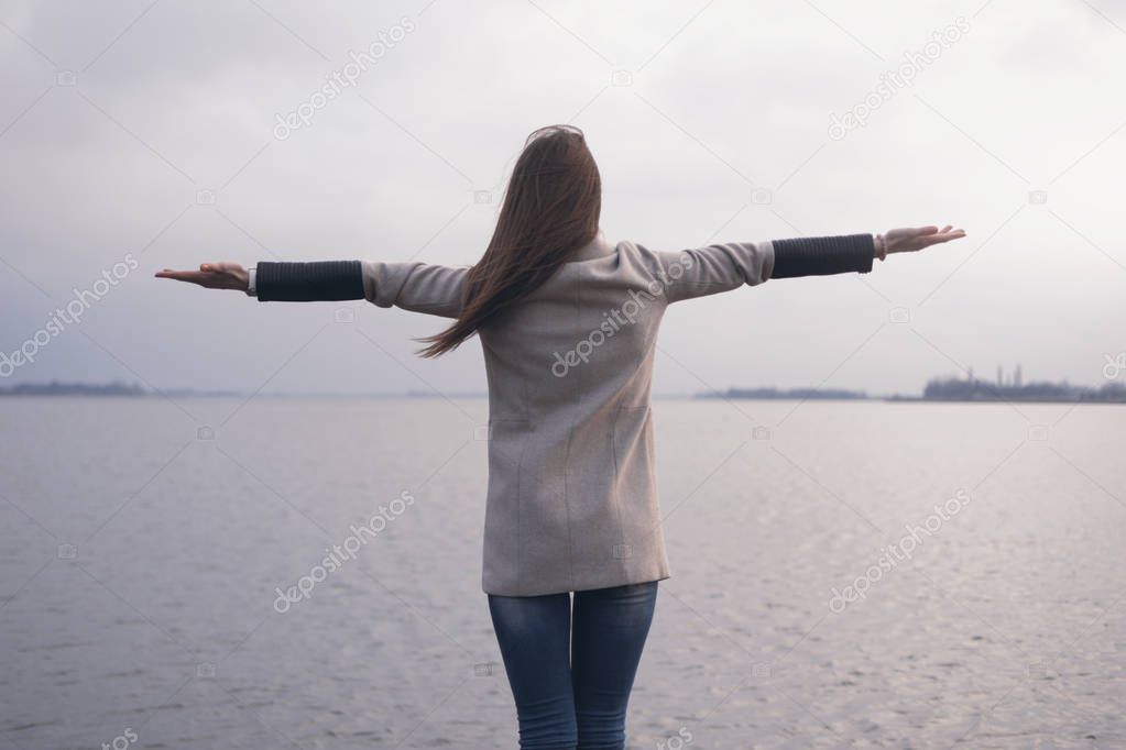 a rear view girl with her arms spread to the lake/Rear view  an adult girl with long brown hair and widespread hands standing on the shores of the lake