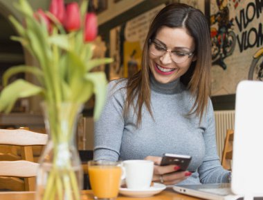 açık bir dizüstü bilgisayar önünde otururken bir akıllı telefon kullanarak genç iş kadını. kahve dükkanında bir cep telefonu ve dizüstü bilgisayar kullanarak gülümseyen kadın