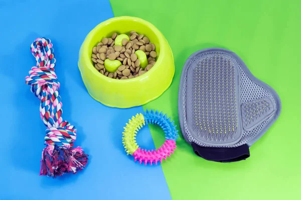 Pet supplies about bowl with dry food, rubber toys, glove brush and rope on blue and green background.