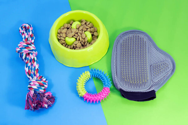 Pet supplies about bowl with dry food, rubber toys, glove brush and rope on blue and green background.