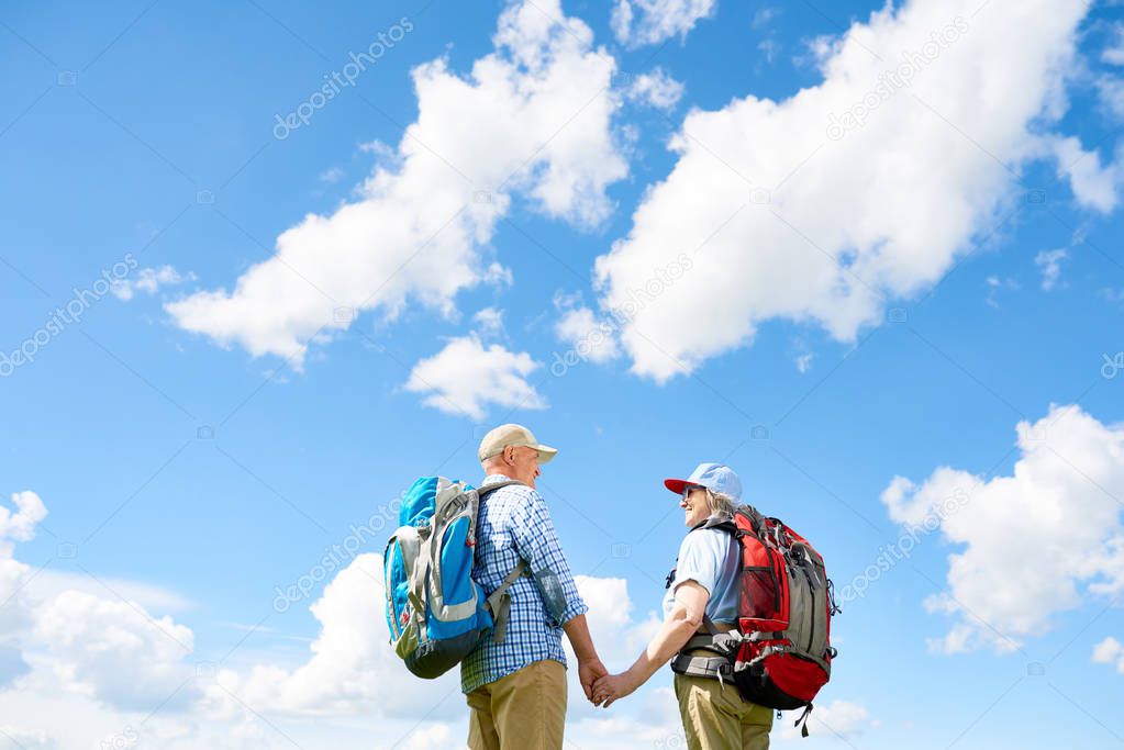 Back view portrait of active senior couple enjoying hiking trip together standing with big backpacks holding hands against background of clear blue sky