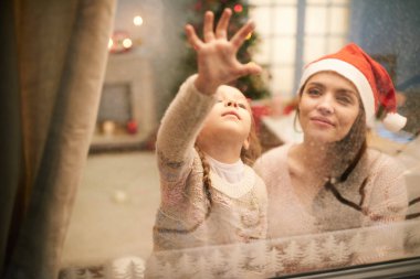 Rahat oturma odası bir araya süre güzel anne ile eğleniyor örme kazak giyen sevimli küçük kız Noel Kutlamaları ve çizim resim donmuş penceresinde için dekore edilmiş.