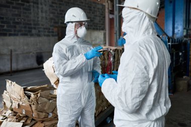 İki işçi biohazard takım elbise arka planda geri dönüşümlü karton yığınlarının modern atık işleme tesisi, endüstriyel depoda iletişim giyen portresi