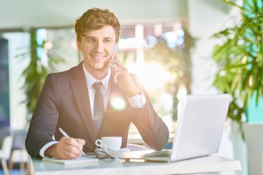Telefonla konuşma ve güneşli kafede kahve molası sırasında çalışırken gülümseyen yakışıklı genç işadamı portresi