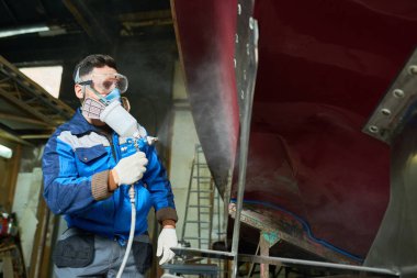 Low angle portrait of unrecognizable worker wearing protective mask painting boat using paint sprayer in yacht workshop, copy space clipart