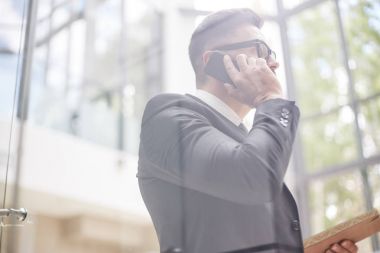 Profil görünümü klasik giyiyor emin orta yaşlı girişimci meslektaşı için smartphone duran geniş ofis lobisinde konuşurken takım elbise, cam duvardan görüntülemek