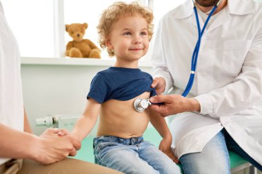 Doktor, cesur ve gülümseyerek, seyir ziyaret sevimli küçük çocuk portresi holding anneler çocuk doktoru kalp atışına stetoskop ile dinleme sırasında el