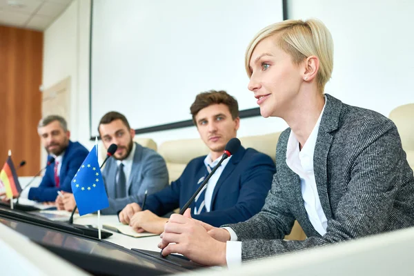 Portrait of several business people sitting in row participating in political debate during press conference focus on young woman answering media questions speaking to microphone