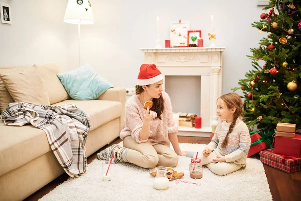 Spending winter evening at home: pretty young woman wearing Santa hat and her little daughter gathered together by fireplace, chatting with each other and enjoying hot chocolate and homemade cookies