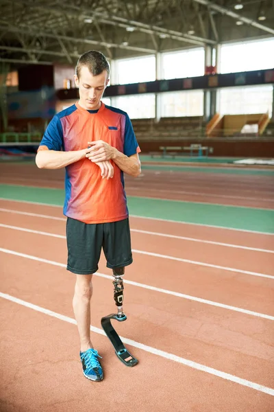 Full length portrait of young amputee athlete checking fitness activity tracker standing on running track in indoor stadium