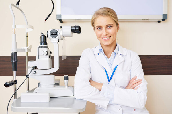 Portrait of blonde female ophthalmologist sitting at slit lamp machine while posing confidently with arms crossed and smiling happily