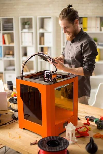 Portrait of contemporary young man enjoying work with 3D printer in modern design studio, using digital tablet and smiling