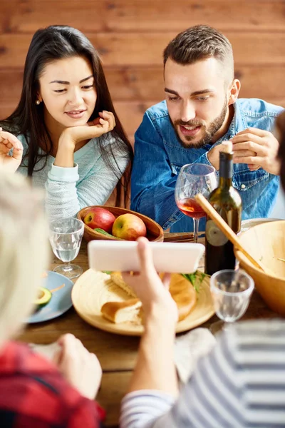 年轻的亚洲妇女和现代男人的肖像看着智能手机屏幕讨论照片 而与朋友坐在大餐桌上的食物 — 图库照片