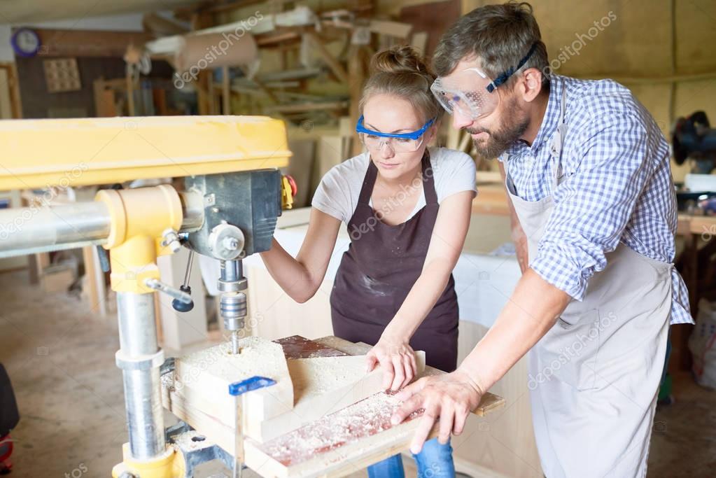 Portrait of two carpenters, man and woman, operating drilling machine making furniture in woodworking shop