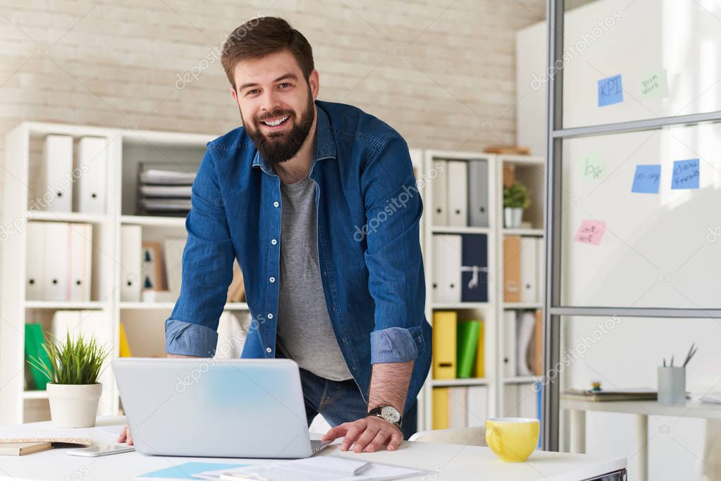 Portrait of modern bearded businessman in casual wear smiling at camera while working with laptop in modern office