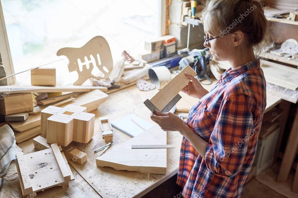 High angle portrait of modern female carpenter measuring wooden part making furniture over working table in workshop