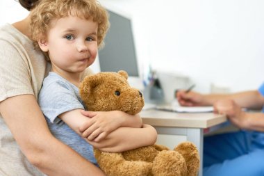 Şirin çocuk anneler üzerinde oturan portresi peluş ayıcık sarılmayı doktor ofisinde tur ve kameraya, seyir alanı Kopyala