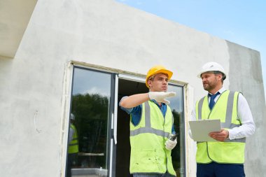 Yakışıklı genç Foreman'ın sakallı orta yaşlı yatırımcı, arka planda bina bitmemiş ile başarılı iş sonuçlarını tartışıyor işçisi ve yansıtıcı ceket