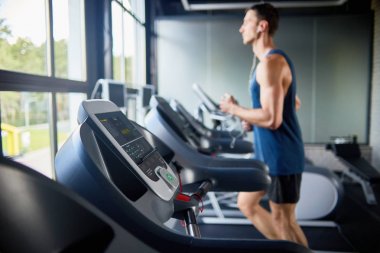 Pencere, odak satır makinelerin üzerinde ön planda tarafından modern spor salonunda koşu bandı üzerinde çalışan genç adam yan görünüm