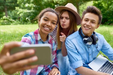 Çok etnik gruptan oluşan grup üç öğrenci selfie açık havada, yeşil çimenlerin üzerine alarak smartphone kameraya görünümlü yapması