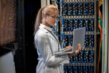 Yan görünüm portre sarışın kadın bilim adamının server dolapları tarafından ayakta ve dizüstü bilgisayar kullanarak süper bilgisayarla çalışma önlük giyiyor