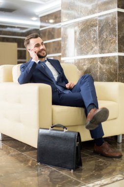 Telefonla konuşma ve deri koltukta oturan otel lobisinde beklerken gülümseyen yakışıklı sakallı işadamı portresi tam uzunlukta