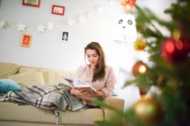 Genç ve güzel kadın portresi tam uzunlukta rahat koltukta oturan ve roman, rahat oturma odası arka plan üzerinde Noel kutlaması için dekore edilmiş iç okuma ekose kaplı